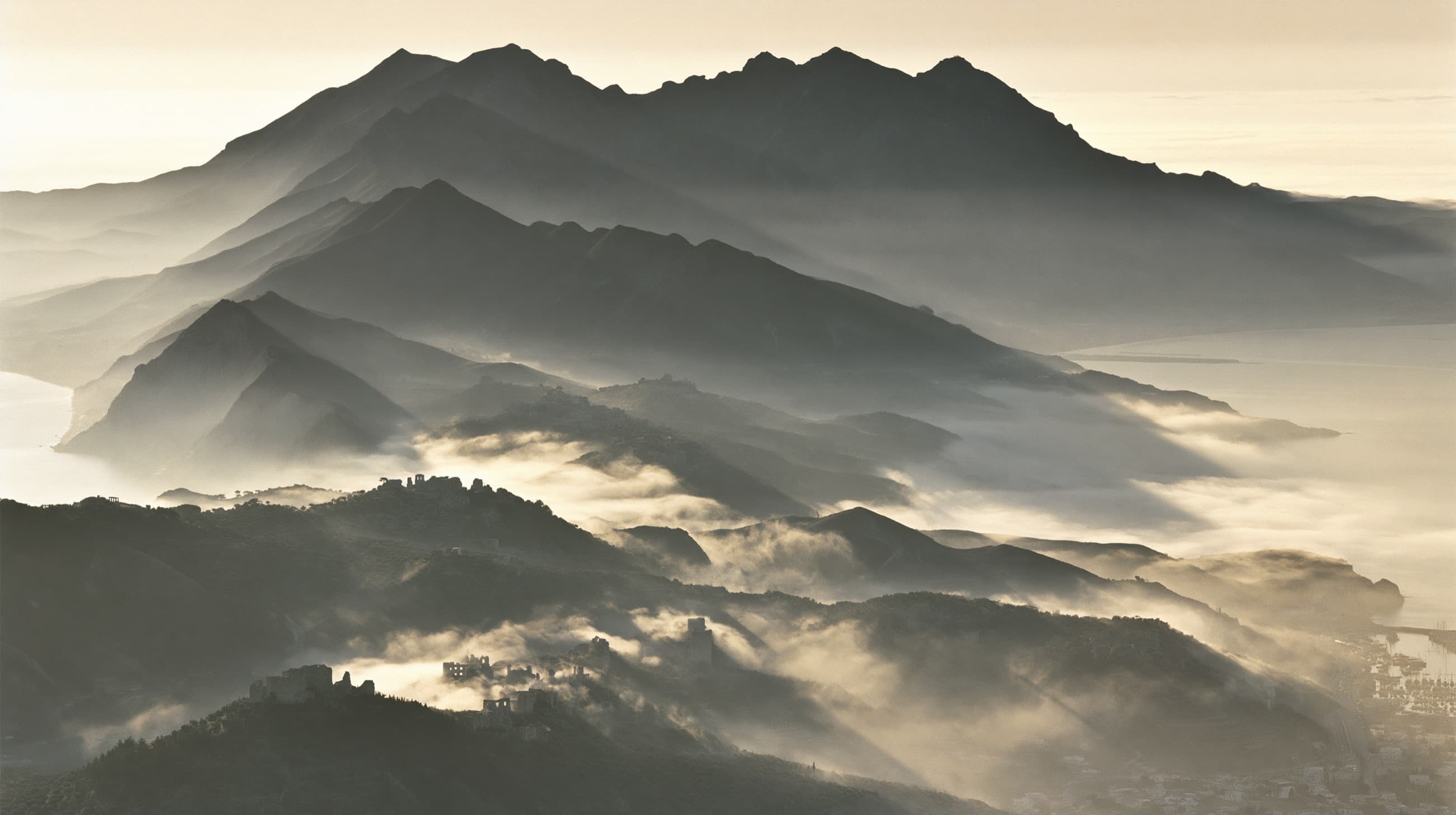 Mountain ridgelines at dawn seen from high altitude, hilltop villages and ruins surfacing through banks of fog, a harbor town far below.